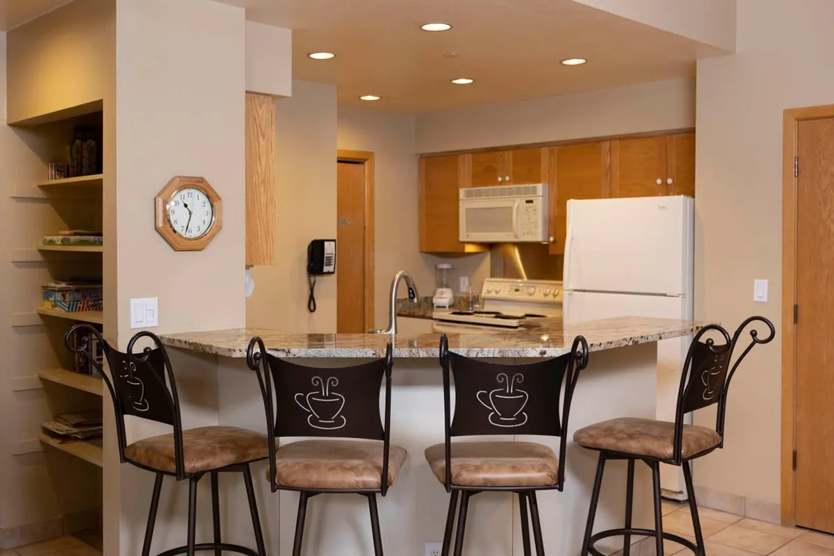 A view of the kitchen with a granite counter, bar stools, modern appliances, and a shelf lined with books. The layout encourages both cooking and social interaction, with warm lighting offering a welcoming ambiance.