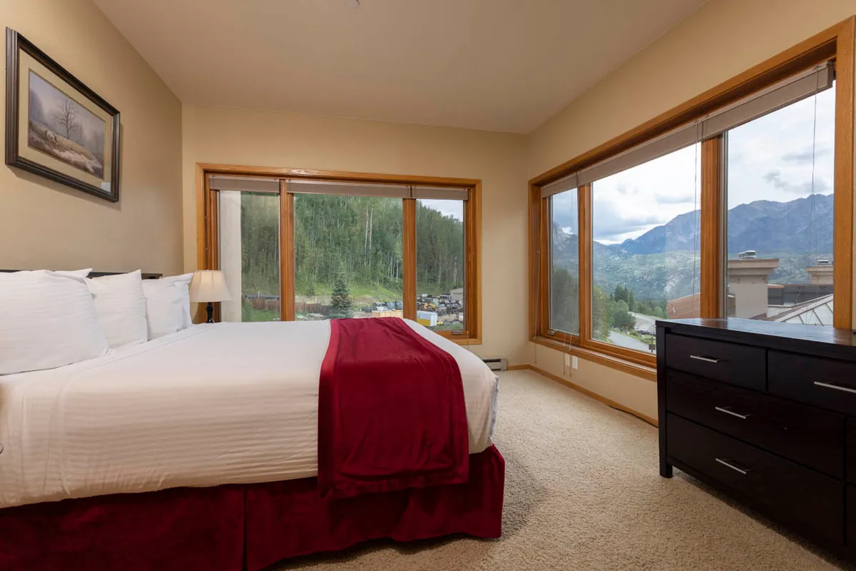 A peaceful bedroom scene featuring a large window that frames stunning mountain views, a cozy bed with a red blanket, and a dark wooden dresser. Soft natural light fills the space, creating a welcoming retreat ideal for relaxation and unwinding after an active day.