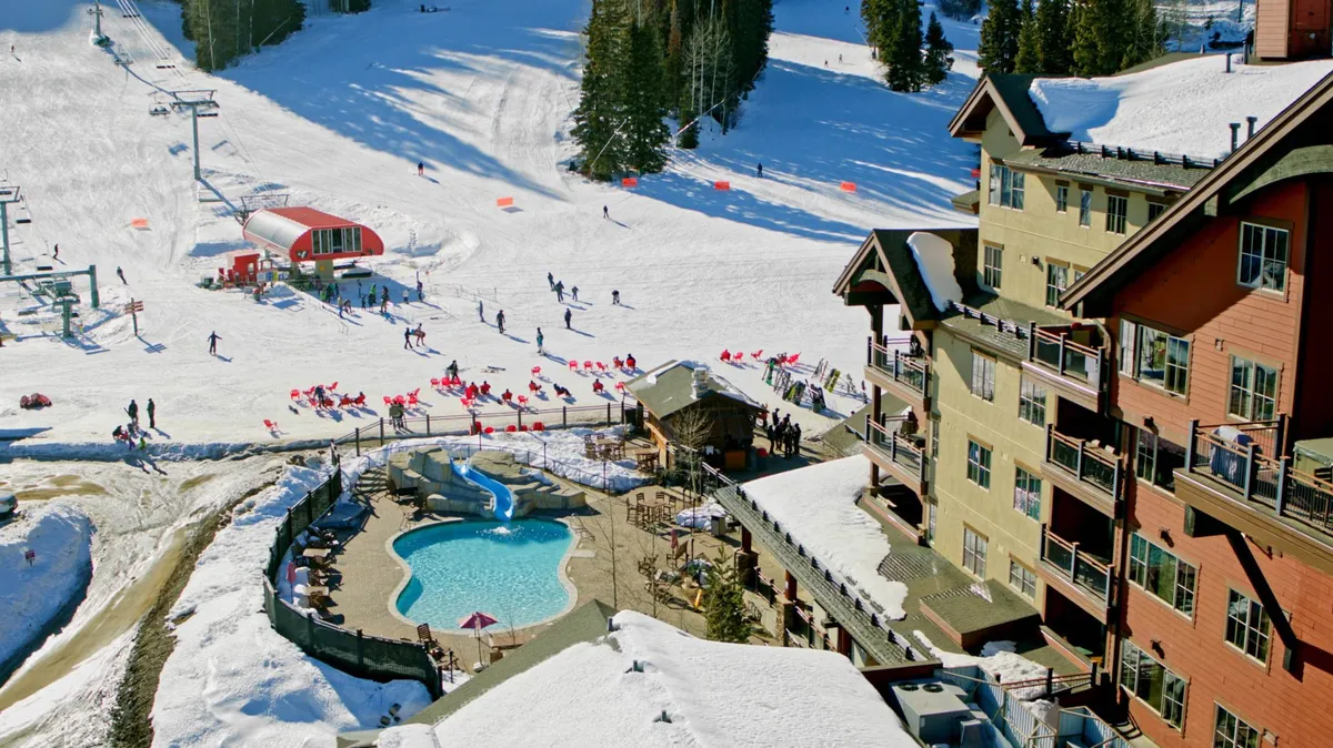 A lively scene showing ski slopes, a swimming pool, skiers and snowboarders in action, and lodge buildings. The pool and lodge are clearly visible, creating a sense of energetic resort life and offering a glimpse of the variety of amenities available.