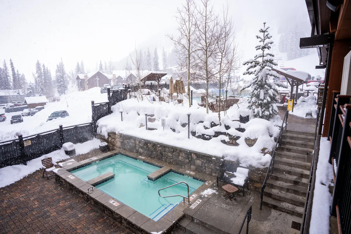 An outdoor hot tub surrounded by snow, with steps leading up and a tranquil winter landscape in the background. The hot tub area exudes peace and relaxation, with the snowy environment enhancing the sense of après-ski indulgence and comfort.