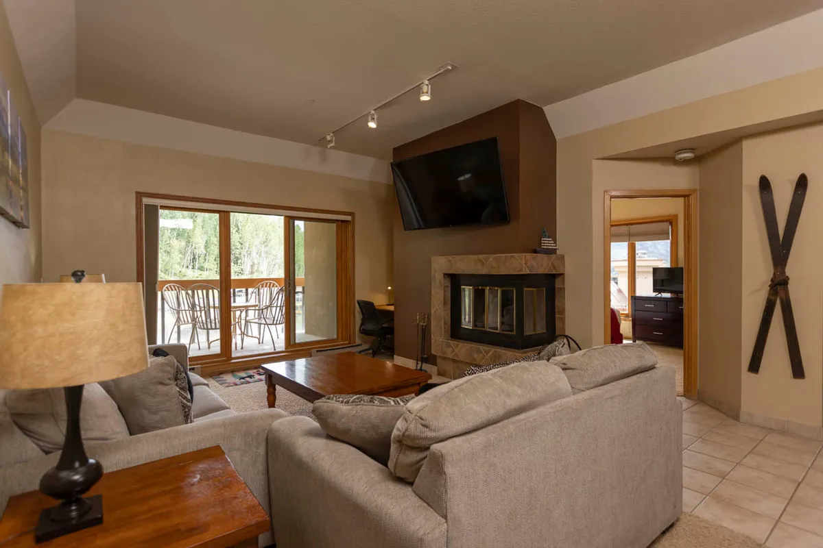 The hero image showcases a cozy living area with a crackling fireplace, inviting sofa, large windows with mountain views, a modern TV, and a dining space—all bathed in warm, natural light. The scene radiates comfort and relaxation, perfect for après-ski gatherings and soaking in the surrounding alpine beauty.