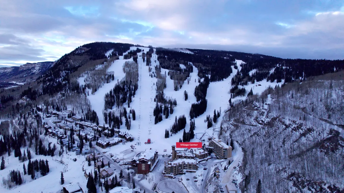 This image captures the snowy ski slopes adjacent to the Village Center, flanked by picturesque pine trees and dramatic mountain scenery. The village buildings are visible at the base, emphasizing the proximity between lodging and the action on the slopes.