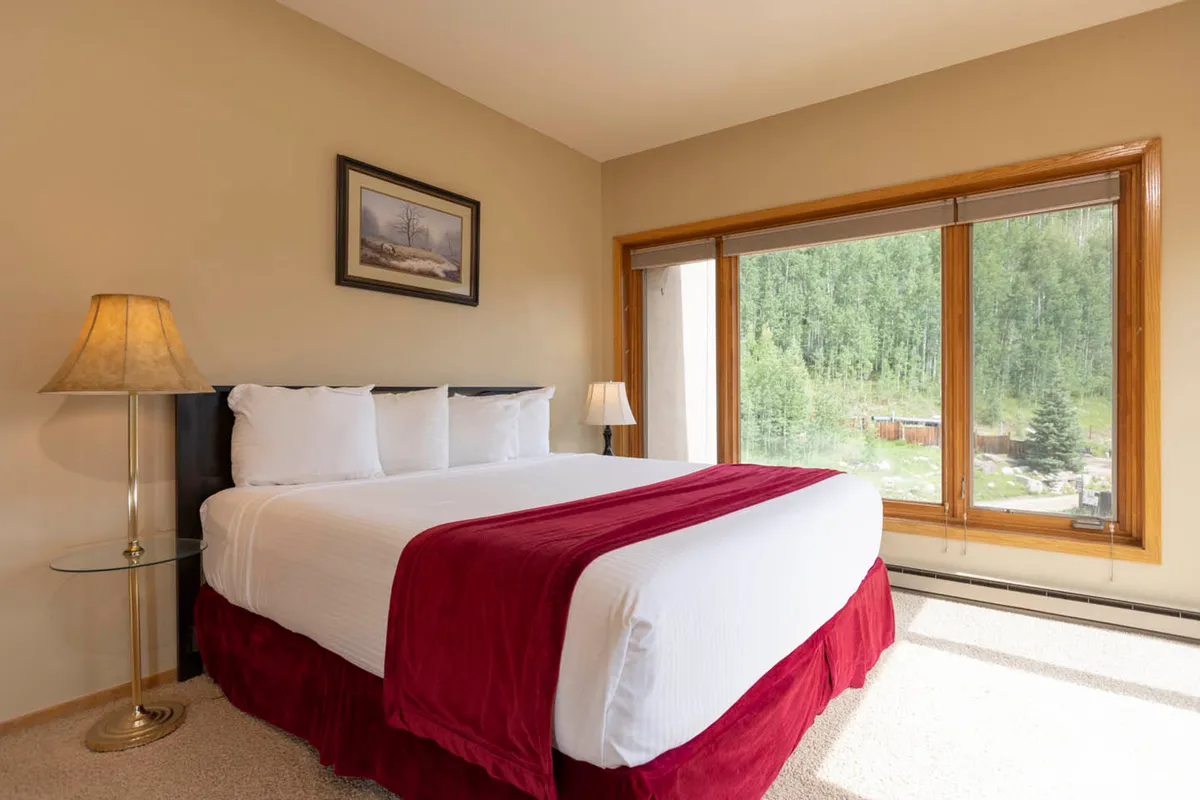 A second serene bedroom with a comfortable bed, large window letting in natural light, bedside lamp, and tasteful artwork on the wall. The space feels calm and private, perfect for recharging after a day on the slopes.