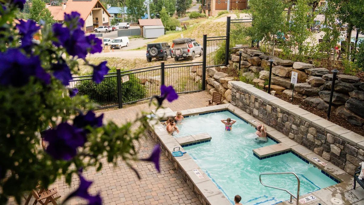 A sunlit pool area with an outdoor hot tub bordered by vibrant flowers and panoramic mountain views. The inviting scene suggests relaxation and socializing, with the surrounding scenery reinforcing the resort setting.